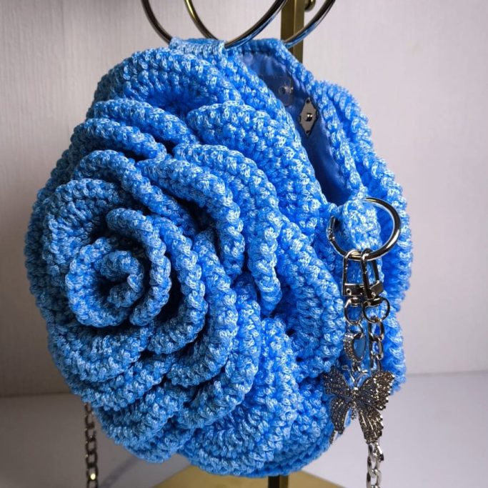 Modell: „Blue Rose“ • Referenz: CF-CEC07 • Beschreibung: Kunstvoll gehäkelte Rosen-Optik mit plastischen Blütenblättern. Ein absoluter Hingucker für besondere Anlässe. • Farbe: Lebendiges, meliertes Himmelblau. • Details: Zwei runde, silberne Metall-Handgriffe; abnehmbare Silberkette mit gehäkeltem Schulterschutz. • Maße: Ø 25 cm (Durchmesser) • Preis: 189 € (zzgl. Versandkosten)